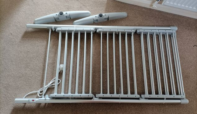 The Swan 3 Tier Heater Airer folded up and laying on biscuit-coloured carpet. The two grey plastic feet of the airer are sitting next to it