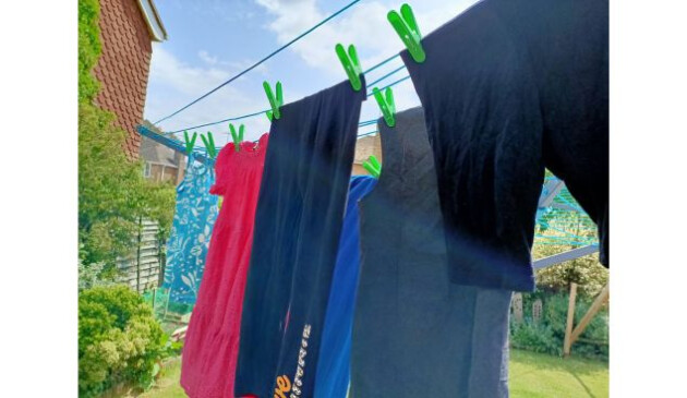 Children's clothes, including a pink dress, grey school pinafore, black leggings and black cycling shorts, hanging from a rotary washing line with green clothes pegs.