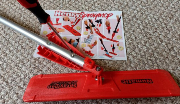 A close up of the Henry Spray Mop floorhead which is laying on a beige carpet next to the instructions. A red and black screwdriver is coming in from the upper left of the picture to tighten a screw on the bottom of the mop handle