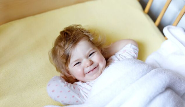 a little girl making a cheeky face, tucked up in her cot