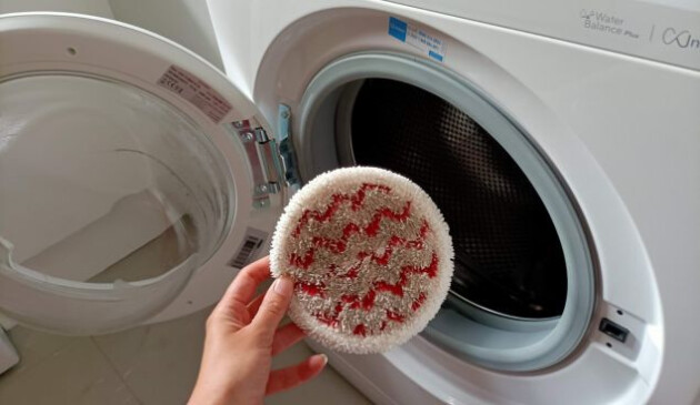 A woman's hand coming from the bottom left of the picture holding a circular microfibre pad which is very dirty. A white washing machine with an open door sits in the background