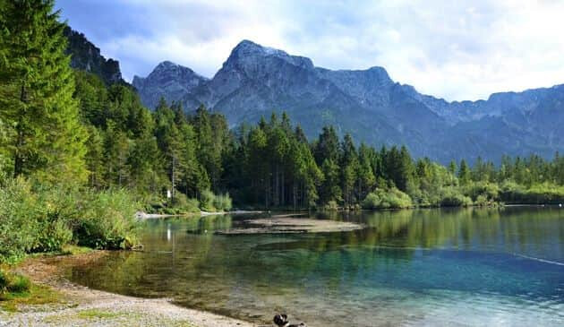 Light blue lake with green grass and lush pine trees with mountains and cloudy sky in the background