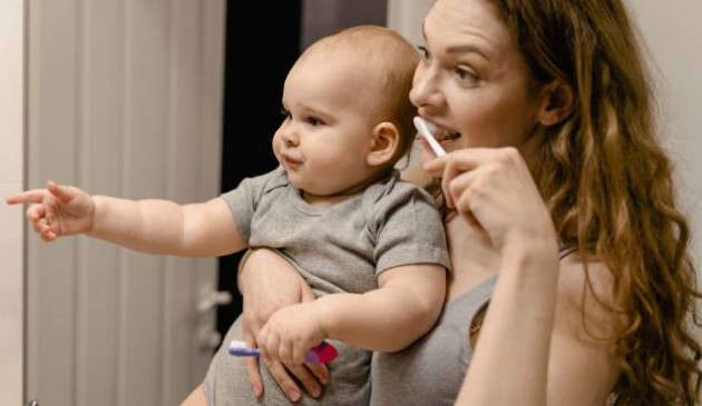 mother holding baby and showing them how to brush teeth in bathroom mirror