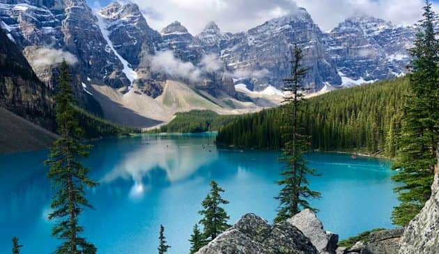 Cloudy skies with snow top ountains overlooking lush green pine trees and a large, bright blue lake