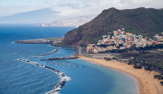 view from above of the blue water and sand with green mountains, cloudy skies and a cluster of small buildings facing the water.