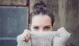 A brunette woman with her hair in a bun and blue eyes. She is wearing a cream jumper and has pulled the collar up over her nose. She is looking straight ahead.