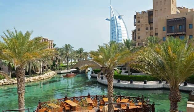 Manmade blueish-green lake in the middle of a patio with tall palm trees, a rustic brown building and a hotel that is shaped like a sailboat in the background