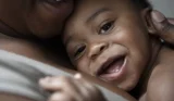 A child smiles at the camera while hugged to his mum's chest, she's out of shot