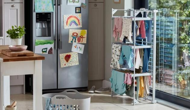 A picture of a heated airer by a kitchen window. In the background is a fridge with children's pictures on.