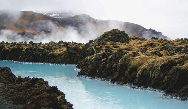 Stream of blue water between two grassy bank with steam and mountains in the background