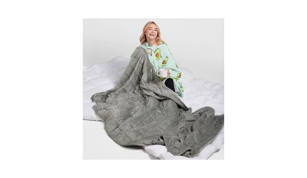 A blonde, smiling woman wearing an over sized green hoodie with avocado print. She has a large, grey blanket on her lap.
