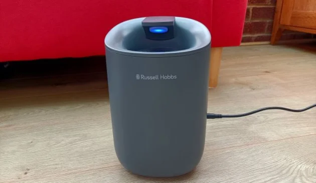 A small, grey Russell Hobbs dehumidifier with a blue light on and black power lead, standing on a wooden floor with a bright red sofa in the background