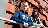 Two young people in athletic wear lean against a railing against a backdrop of geometric terracotta-colored architecture.
