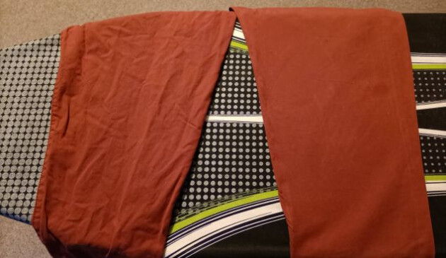 A pair of terracotta-coloured trousers laid out on a black, grey, white and lime green ironing board. One trouser leg is wrinkled and creased, the other is smooth.