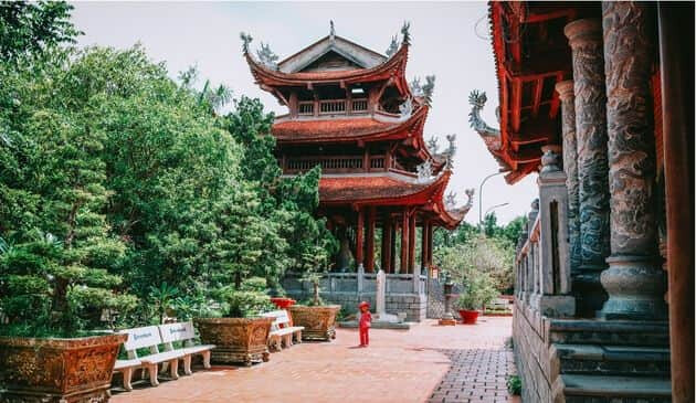 Traditional red Vietnamese building with a three storey roof with green forestry to the left of it and another traditional building with pillars to the front of it.